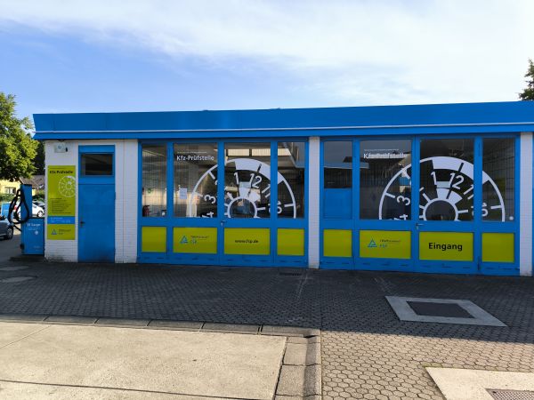 Außenansicht einer Kfz-Prüfstelle mit blauer und gelber Beschilderung, großen TÜV-Plaketten als Fensterdekor, einer Eingangstür und einem Staubsaugerautomaten links.