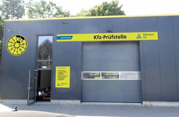 Modernes, graues Gebäude mit gelbem Schild "Kfz-Prüfstelle", geöffnetem Eingang und großer Torzufahrt; links oben eine gelbe Plakette, Bäume im Hintergrund.